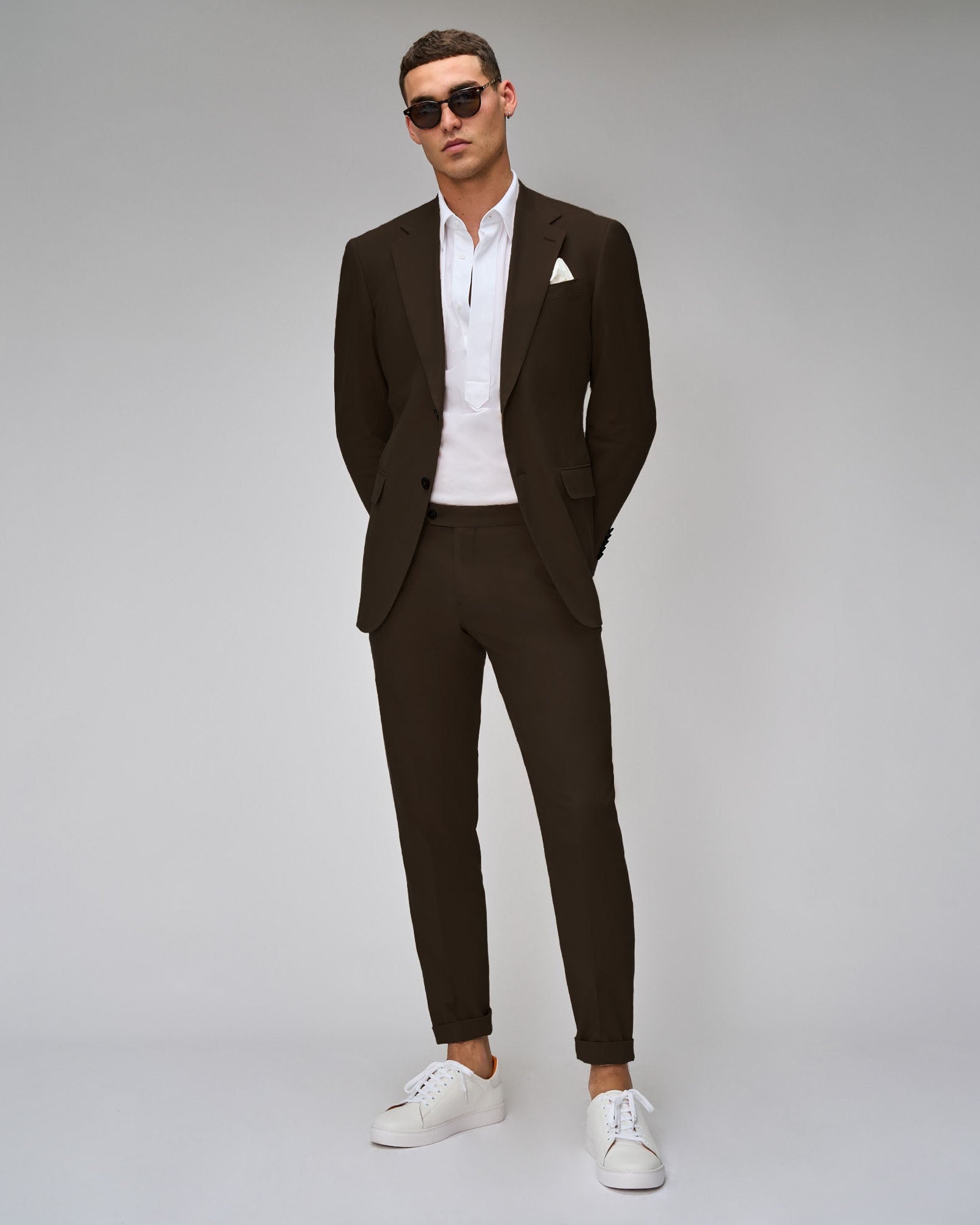 A man standing against a white background, wearing a brown suit with a shirt and tie, paired with white sneakers.