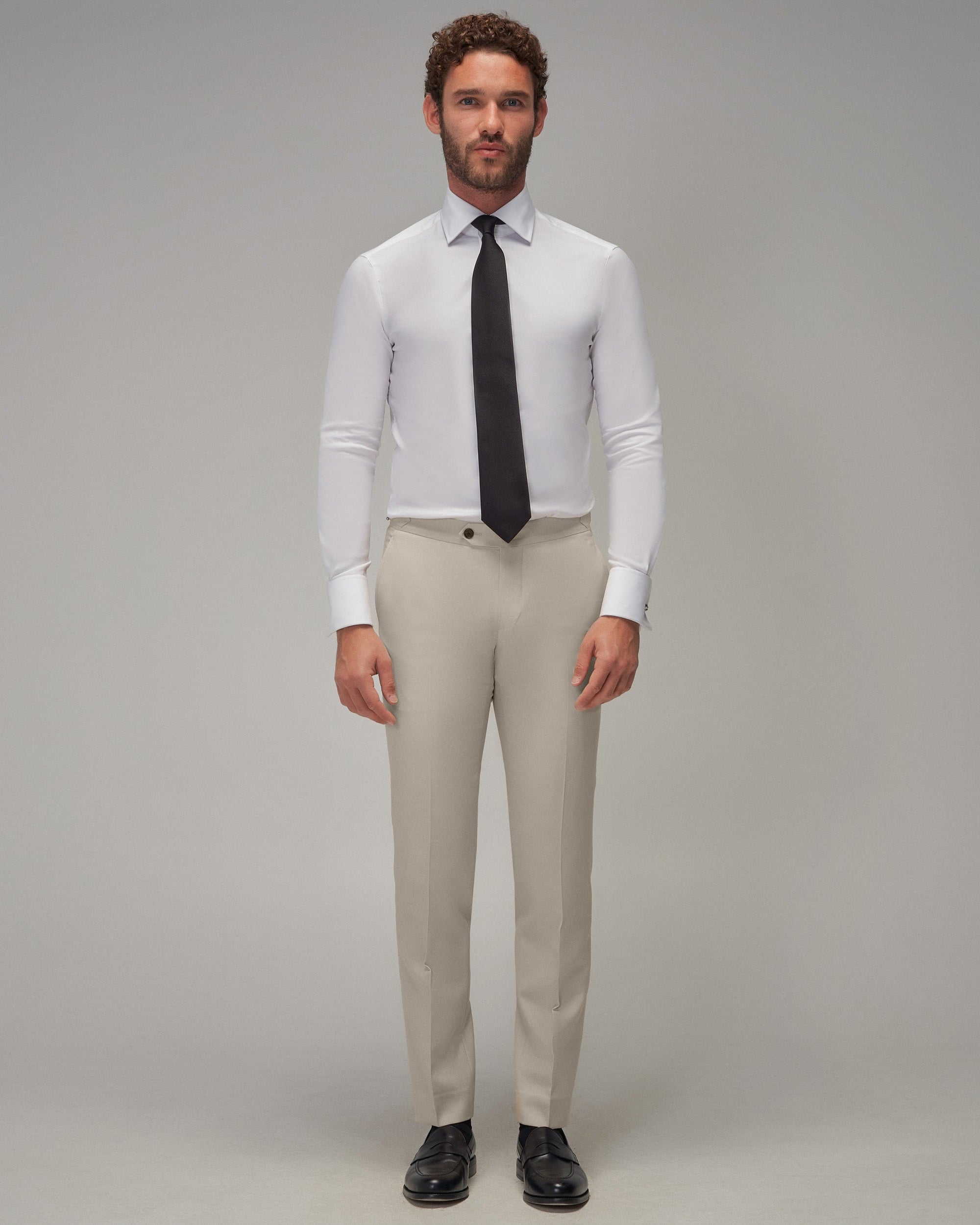 Man wearing a white dress shirt, black tie, beige pants, and black shoes on a gray background