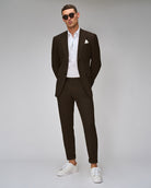 A man standing against a white background, wearing a brown suit with a shirt and tie, paired with white sneakers.