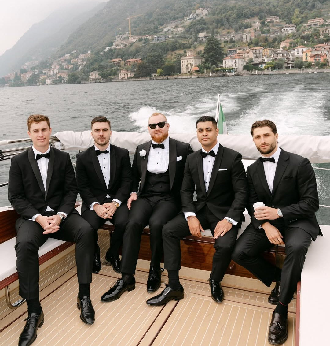 Five men in tuxedos suits sitting on a boat with a scenic mountain backdrop.